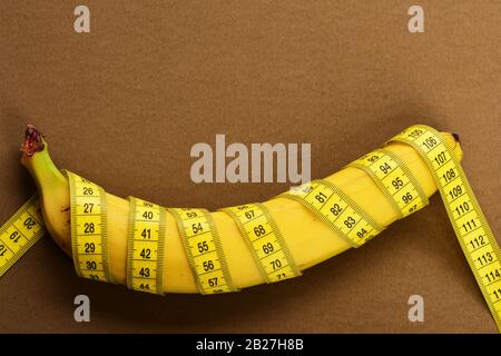 Rolled centimeter ruler spinned around fresh fruit. Banana with yellow tape for measuring figure. Yellow tape around banana isolated on brown background. Bodybuilding and healthy diet concept. Stock Photo