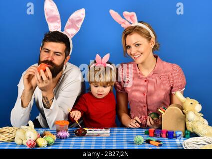 Mother, father and daughter with smiling and curious faces preparing for Easter. Man, woman and kid wearing bunny ears. Family members painting eggs on blue background. Holiday spirit and joy concept. Stock Photo
