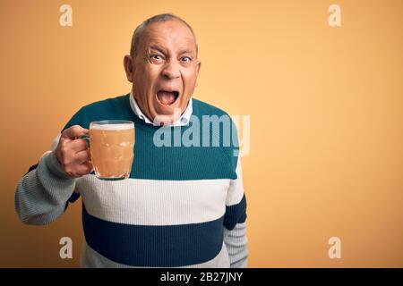 Senior handsome man drinking jar of beer standing over isolated yellow background angry and mad screaming frustrated and furious, shouting with anger. Stock Photo