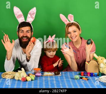 Mother, father and son painting eggs in pink bunny ears. Happy moments and Easter celebration concept. Family members preparing for Easter on green background. Man and woman with happy smiles. Stock Photo