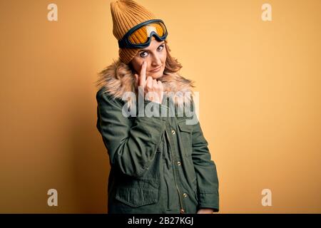 Middle age beautiful blonde skier woman wearing snow sportwear and ski goggles Pointing to the eye watching you gesture, suspicious expression Stock Photo