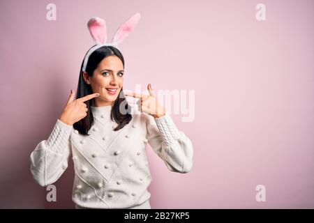 Young caucasian woman wearing cute easter rabbit ears over pink isolated background smiling cheerful showing and pointing with fingers teeth and mouth Stock Photo