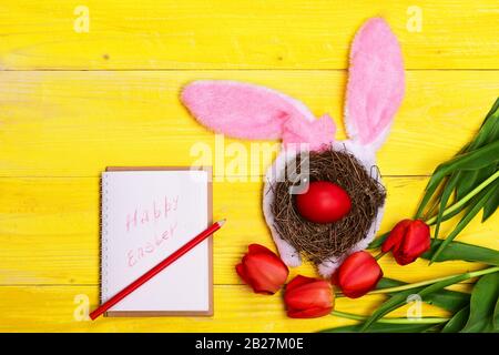Happy holiday concept. Tulips in pink color on yellow wooden background. Bunch of flowers near Easter eggs in nest with bunny ears near notebook with Happy Easter sign. Spring holiday composition Stock Photo