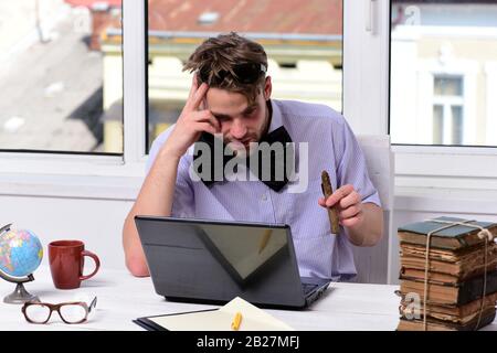 Studying and work online concept. Guy with short hair and bristle wears sunglasses and big black bowtie. Man with troubled face sits by table using laptop. Worker near old books on window background Stock Photo