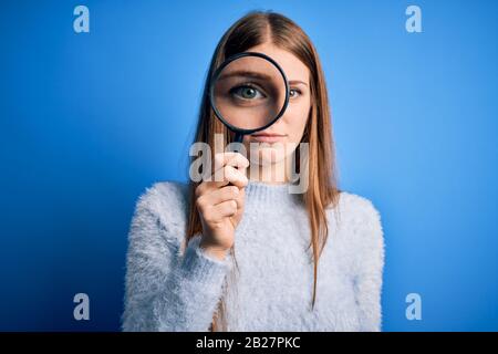 Young beautiful redhead detective woman using magnifying glass over isolated blue background with a confident expression on smart face thinking seriou Stock Photo