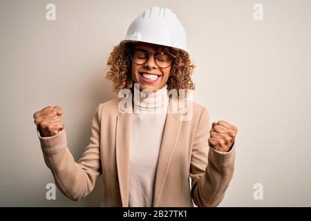 Young african american curly architect woman wearing safety helmet and glasses very happy and excited doing winner gesture with arms raised, smiling a Stock Photo