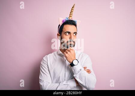 Young handsome man with beard wearing funny unicorn diadem over pink background with hand on chin thinking about question, pensive expression. Smiling Stock Photo