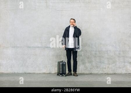Senior man with suitcase standing outdoors and talking on smartphone near a wall Stock Photo