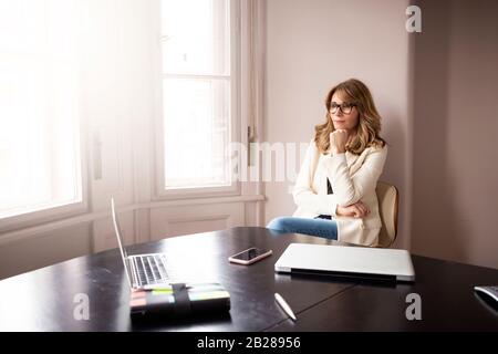 Portrait shot of elegant businesswoman looking thoughtfully while sitting at desk by the window. Stock Photo