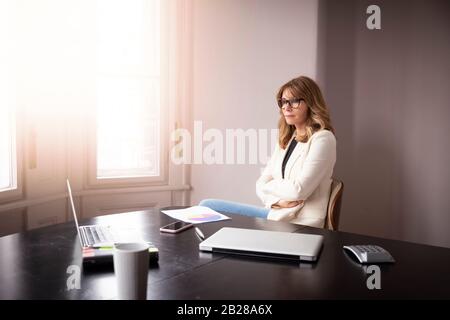 Portrait shot of elegant businesswoman looking thoughtfully while sitting at desk by the window. Stock Photo