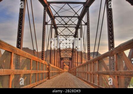 Perspective view a bridge and of its wooden deck walkway with wooden fenced handrails under the metal supporting structures in a warm, rust color Stock Photo