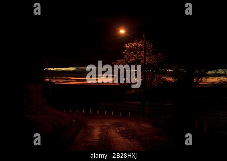 Dark night, lonely road, yellow light from the only street lamp, white poles at the edge,remnants of a beautiful sunset on the road to Tapion Stock Photo