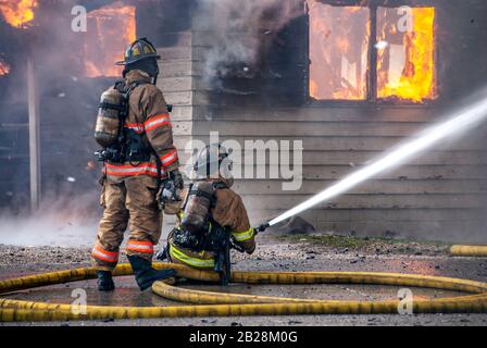 Two Firemen in full protective suits and oxygen tanks working with long yellow water hose spay water onto the burning smoking house engulfed in fire a Stock Photo