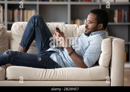 Young african american man lying on cozy sofa, playing games. Stock Photo