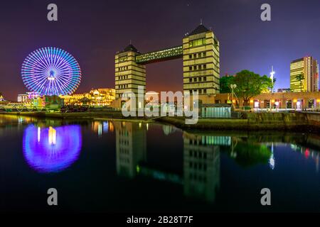 A giant ferris wheel and the buildings in the illuminated waterfront of the bay reflect in the water at night. Cityscape of Yokohama, Japan. Stock Photo