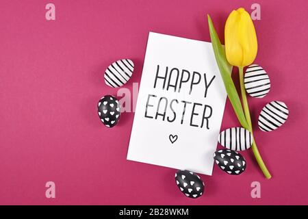 'Happy Easter' text card surrounded by black and white easter eggs and single yellow tulip spring flower on bright pink colored background, flat lay Stock Photo