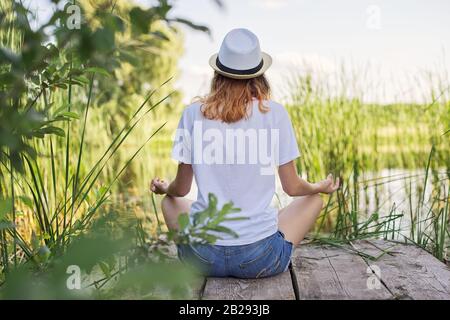 Young woman sitting in lotus position on bridge near water, back view Stock Photo