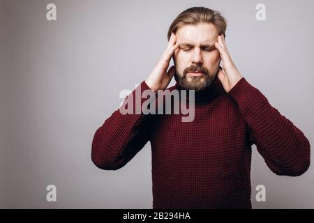 Front view of mature bearded man in knitted sweater touching temples with fingers because of headache. Isolated over grey studio background. Stock Photo