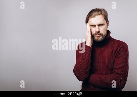 Middle-aged bearded man in claret-colored sweater suffering from headache while posing in studio over grey background. Mature male holding hand on hea Stock Photo