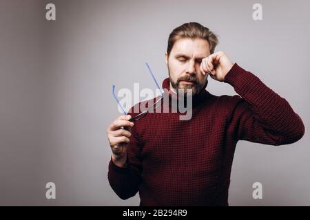 Portrait of bearded man in casual clothing taking off eyeglasses and rubbing his tired eyes. Mature person suffering eye strain while standing over gr Stock Photo
