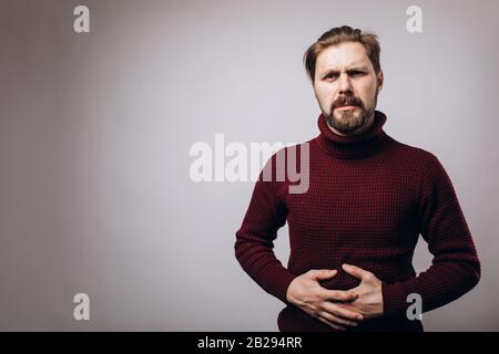 Mature bearded man in warm knitted sweater suffering from severe stomach ache while posing in studio with grey background. Concept of indigestion and Stock Photo