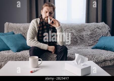 Mature sick man in warm home clothing and knitted scarf sitting on sofa at home near table with medicines. Bearded male using nasal spray to make brea Stock Photo