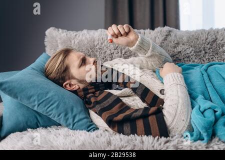 Side view of middle-aged person in warm clothing lying on sofa and covered with blue blanket while looking at high degrees of thermometer. Bearded man Stock Photo