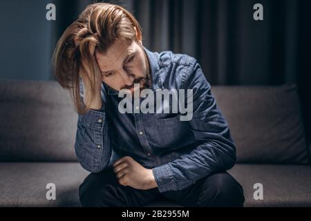 Mature man in casual clothing leaning with elbows on knees and sitting on couch with sad expression. Bearded person feeling depressed at home. Stock Photo