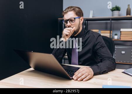 Mature businessman in formal clothing and eyeglasses sitting at modern office and yawning because of feeling tired. Bearded man working after hours to Stock Photo