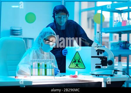 Portrait of two scientists wearing biohazard gear holding box with danger sign while working on research in bio laboratory, copy space Stock Photo