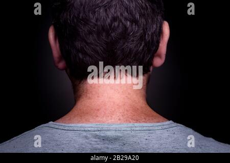 Anonymous caucasian male with short brown hair and grey green shirt on and freckles in his neck seen from behind against a dark background