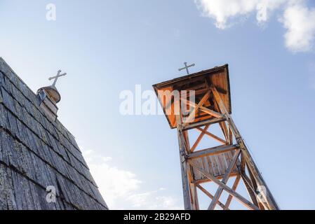 Wooden hut roof built from hay with a cross on top as medieval church in Serbia on a sunny day with a wooden bell tower with brass bell beside it Stock Photo