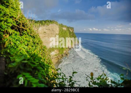 Waves rushing toward a cliff near the Uluwatu temple in Bali with cloudy sky in the background during a late spring afternoon Stock Photo