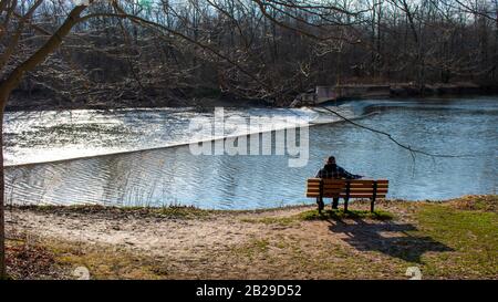 A Lone Man Sitting on a Bench in a Forest Admiring a Waterfall from a Dam and a lake Stock Photo