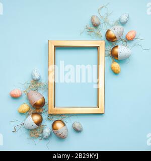 Top view of Easter eggs colored with in different colors arranged around a golden photo frame. Blue background. Copy space. Stock Photo