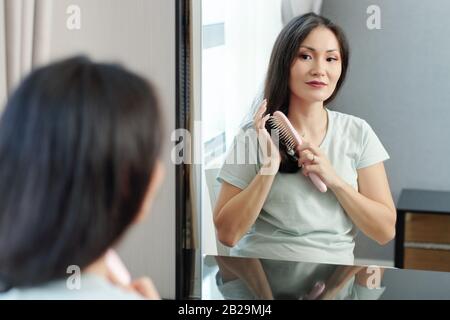 Beautiful young Vietnames woman sitting in front of mirror and enjoying brushing hair Stock Photo