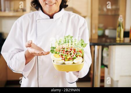 Smiling man in white chef jacket showing bowl of tasty salad he cooked for dinner Stock Photo