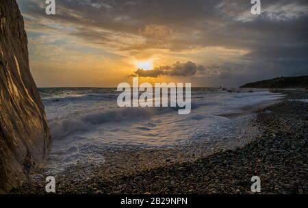 Foamy sea washing by a rocky beach against sunset background. Scene on the Aphrodite's Rock (Petra tou Romiou  or Rock of the Greek) - Cyprus. Stock Photo