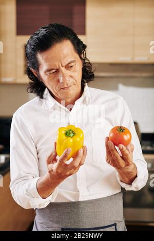 Pensive mature man looking at ripe organic bell pepper and tomato in his hands when standing in kitchen Stock Photo