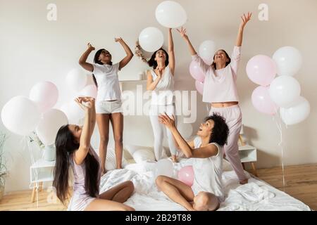 Happy diverse girls playing with balloons in bedroom at party Stock Photo