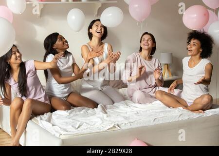 Happy diverse girls playing with balloons, sitting on bed Stock Photo