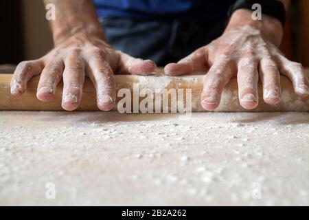 Man's hands are rolling out dough in flour with rolling pin in her home kitchen. Homemade noodle or pasta production by father. Closeup, selective foc Stock Photo