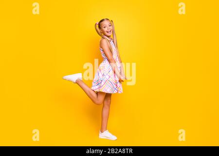 Full length profile photo of beautiful small lady tourist good mood pretty long tails playful wear plaid summer dress white sneakers isolated yellow Stock Photo