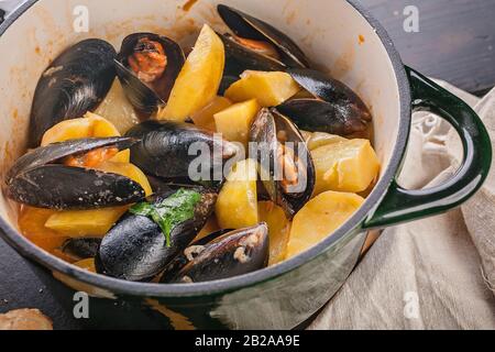 Close-up mussels and fried potatoes in sweet and sour sauce in a saucepan and chabatta on a dark wooden table. Mediterranean traditional cuisine. Stock Photo