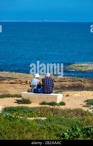 Two old people sitting on concrete bench looking at the ocean, Torrevieja, Costa Blanca, Spain Stock Photo