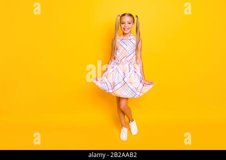 Full length photo of beautiful small lady hold sundress skirt hands good mood pretty long tails wear plaid summer dress white sneakers isolated yellow Stock Photo