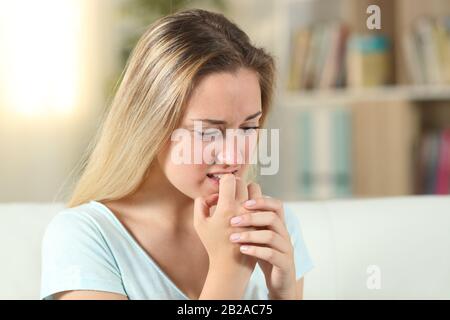 Stressed blonde teenage girl biting her nails sitting on a couch at home Stock Photo