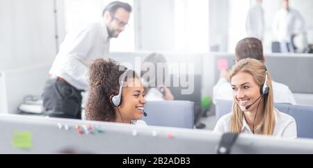 Call center worker accompanied by her team. Smiling customer support operator at work. Young employee working with a headset. Stock Photo
