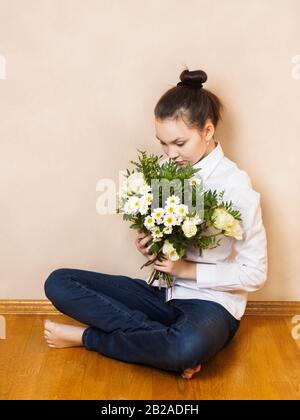 A young girl with a pensive look in a white shirt and jeans sits on a wooden floor with a bouquet of flowers of roses, daisies and green plants Stock Photo