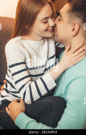 Charming ginger lady with freckles is sitting on a sofa with her husband and kissing him while embracing each other Stock Photo